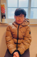 Manuel mit brauner Steppjacke vor Fenster und Kerze sitzt und betet mit geschlossenen Augen.