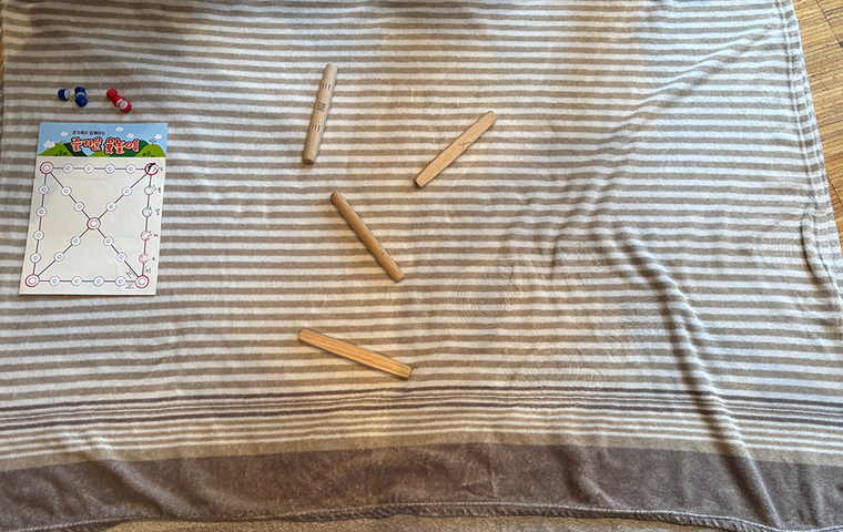 Yut - Spielfeld und 4 Wurfstäbe auf grau gestreifter Decke.