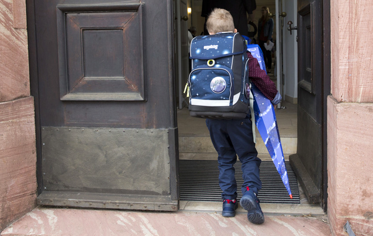 Kind mit Schulranzen und Schultüte geht durch geöffnetes Schultor.